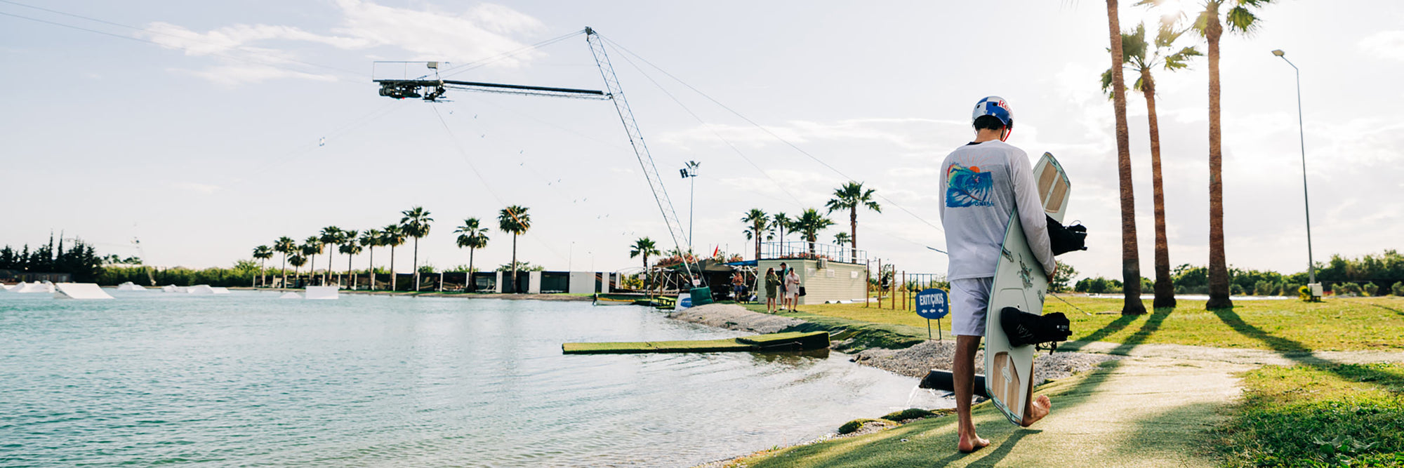 A person in a helmet walks barefoot towards a cable park, holding a wakeboard.