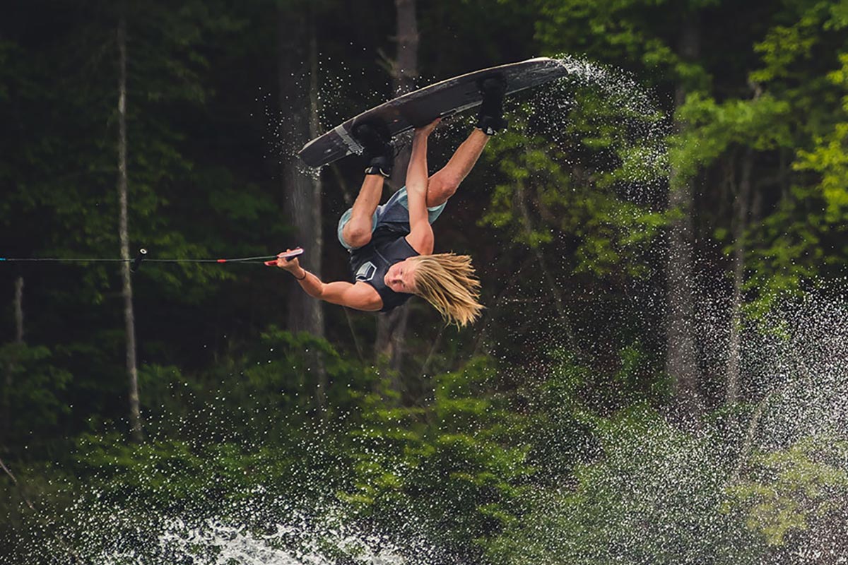 Liquid Force Team Athlete Luca Kidd inverted on a wakeboard with trees in the background on a lake.