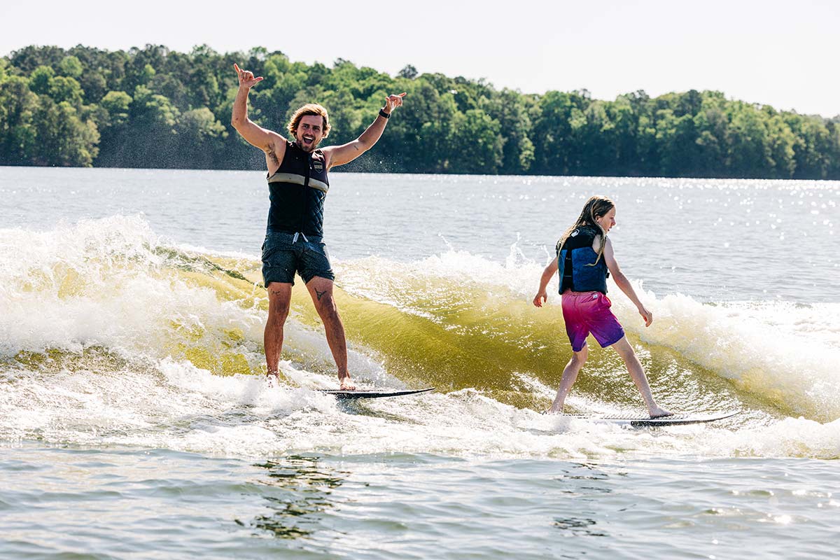 Two people wakesurfing together on a sunny lake, one cheering with arms raised.