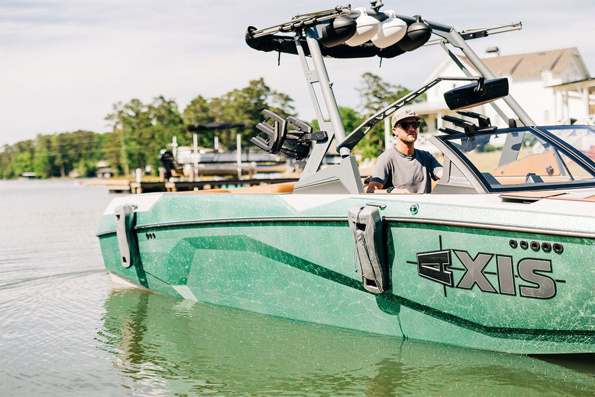 Man piloting a green Axis wake boat near a dock on a sunny day.