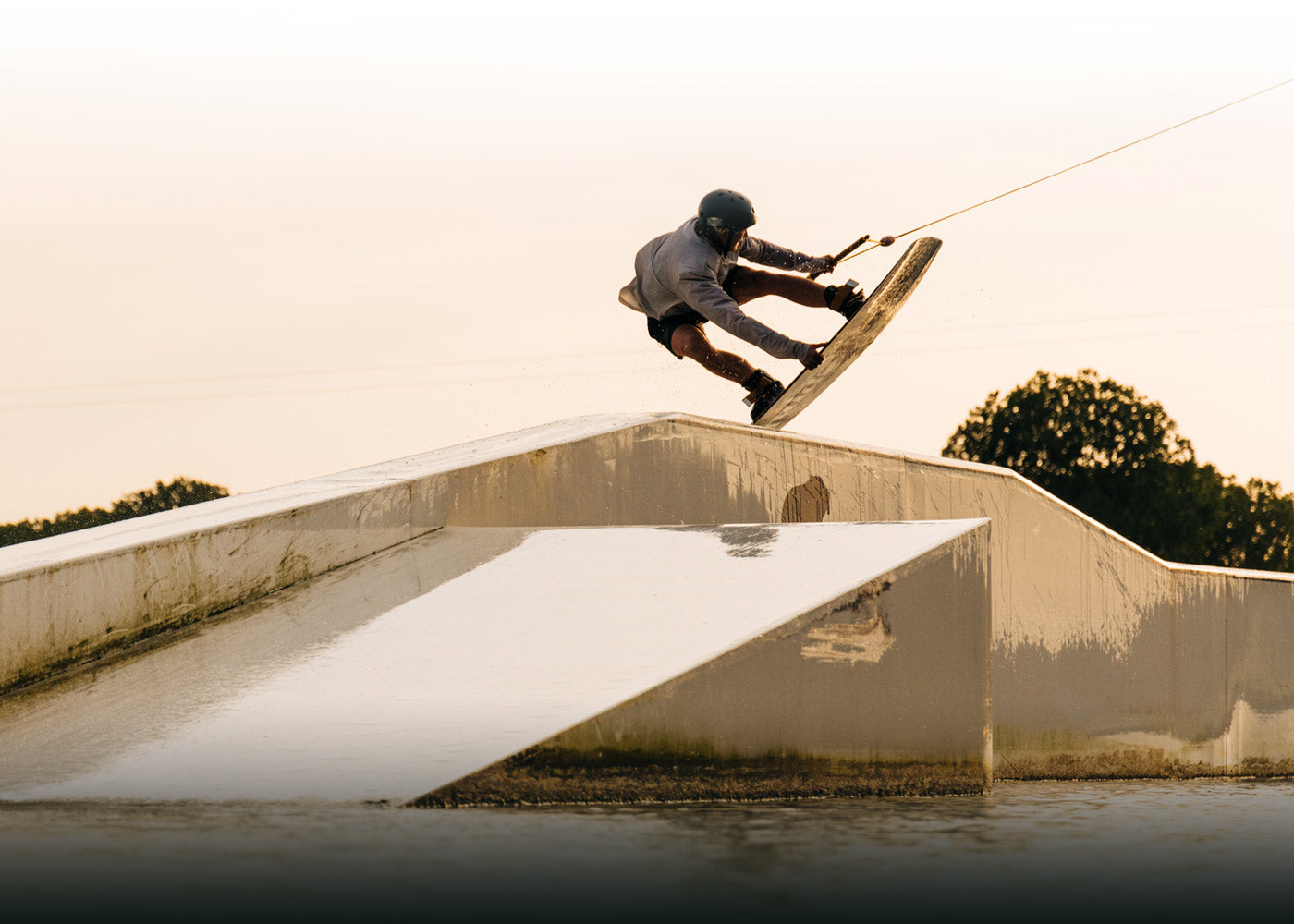 Wakeboarder jumps over a ramp at sunset, with silhouetted trees behind.