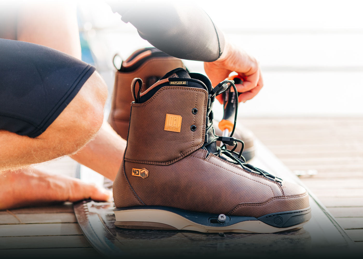 Person tightening laces on a brown wakeboard boot by the water.