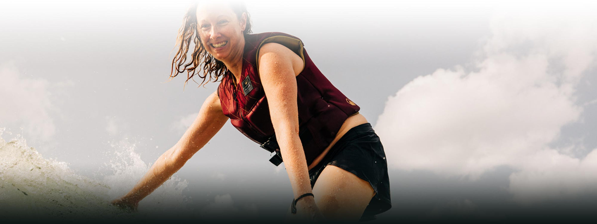 Person surfing, smiling, wearing a red life jacket and black shorts against the cloudy horizon.