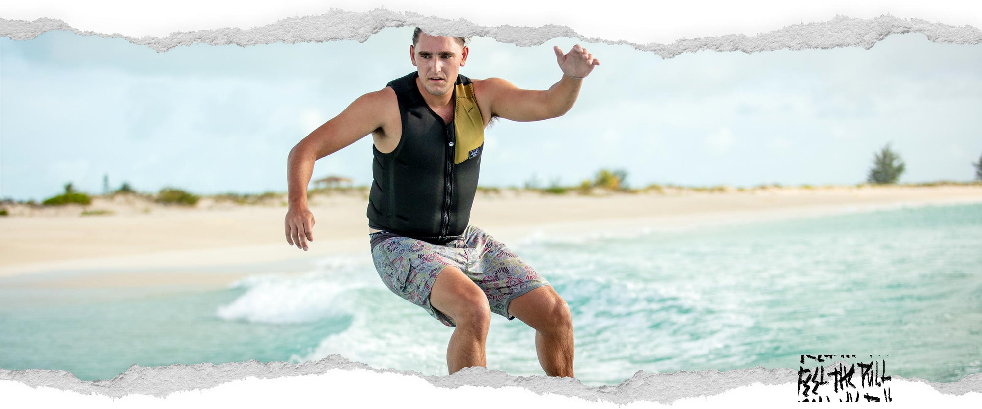 Man wake surfing in a black and olive comp vest with printed boardshorts near a sandy beach.