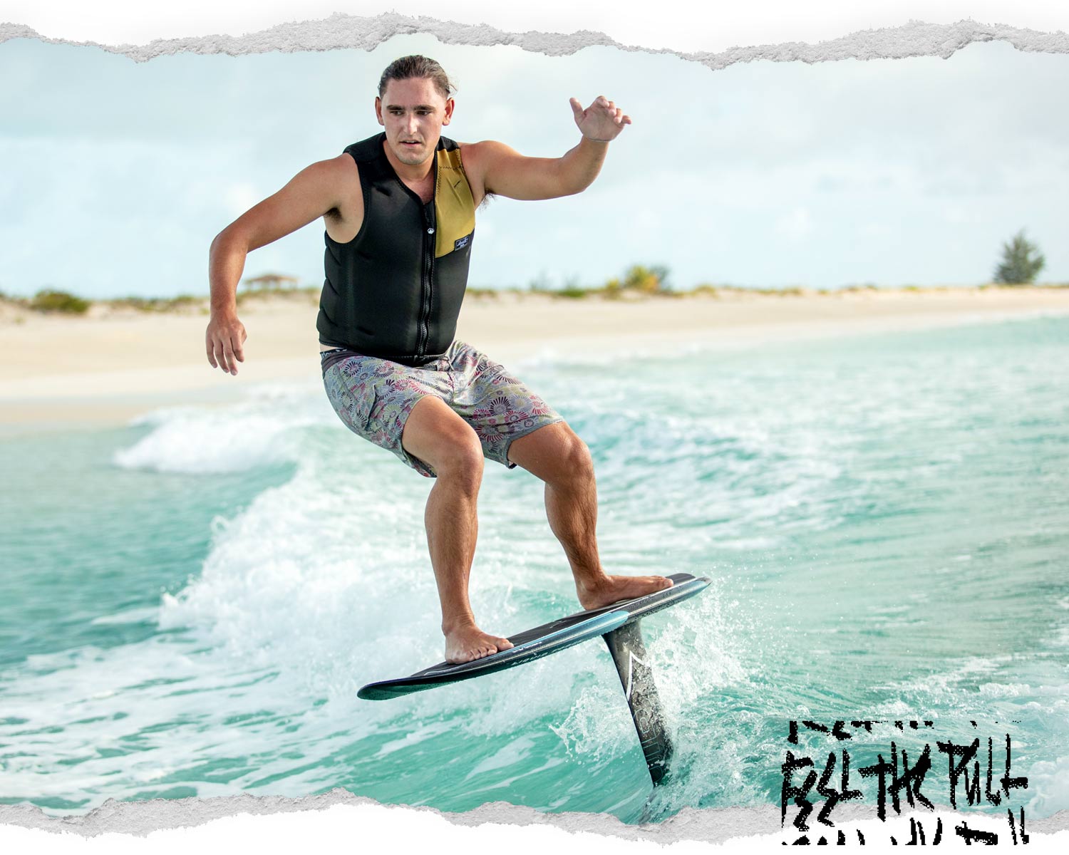 Man wake surfing in a black and olive comp vest with printed boardshorts near a sandy beach.