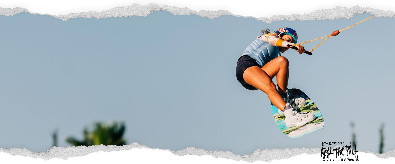 Un rider con chaleco azul claro se lanza al aire en una tabla de wakeboard mientras lleva puesto un casco.