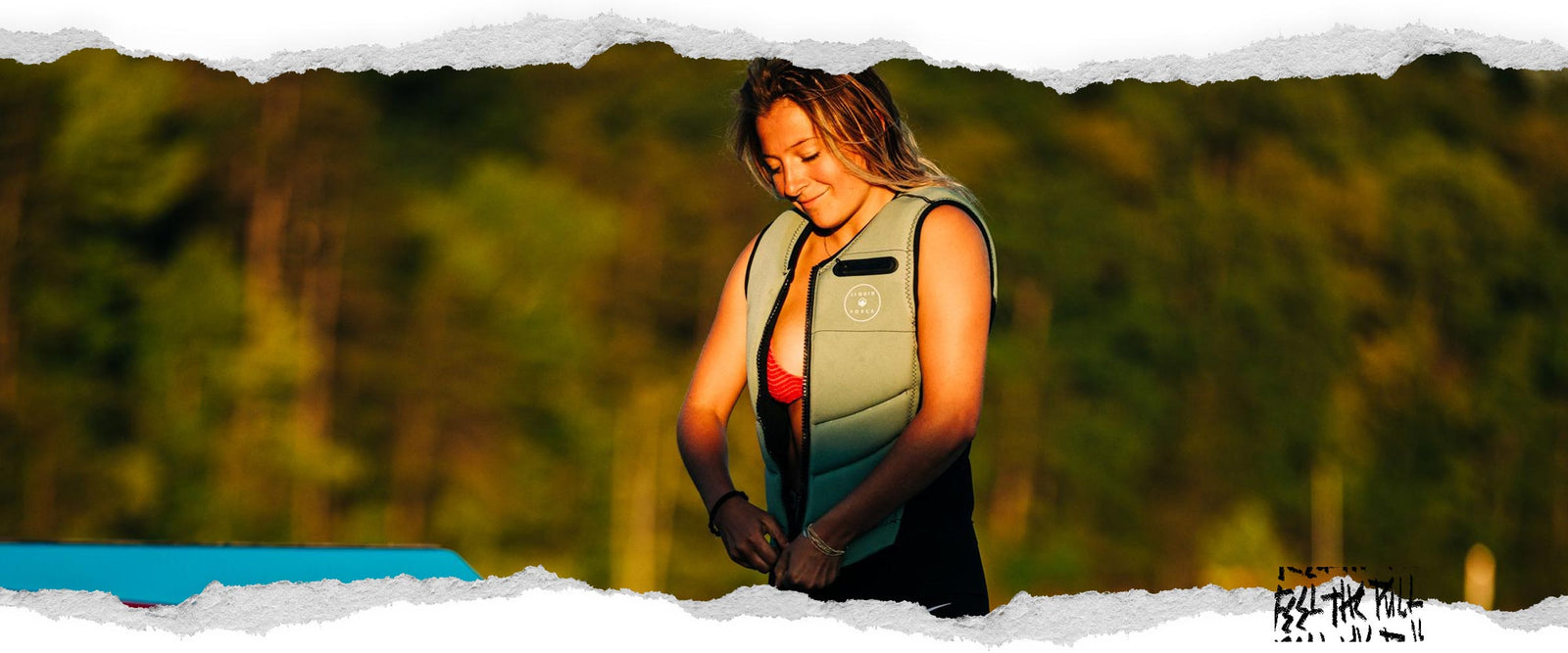 Una mujer se abrocha el chaleco verde Breeze Comp al atardecer junto a un barco azul y rojo.