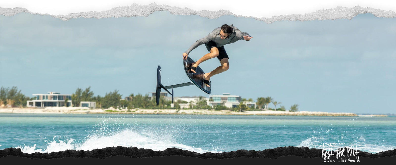 Man in gray shirt jumps a foil board with beach houses in the background.