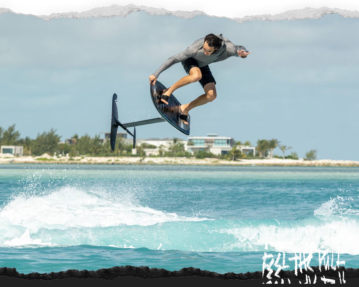 Rider in a gray shirt jumps a foil board over turquoise waves near shore.