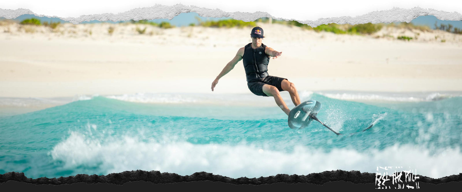 Man in black vest and Red Bull hat balances on a foil board near the beach.