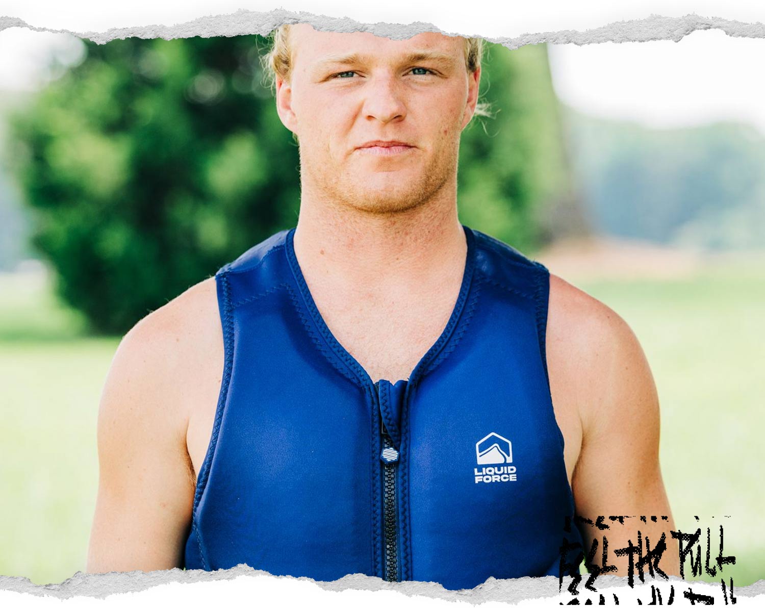 Man in navy Core Comp vest stands outdoors in grass with trees in background.