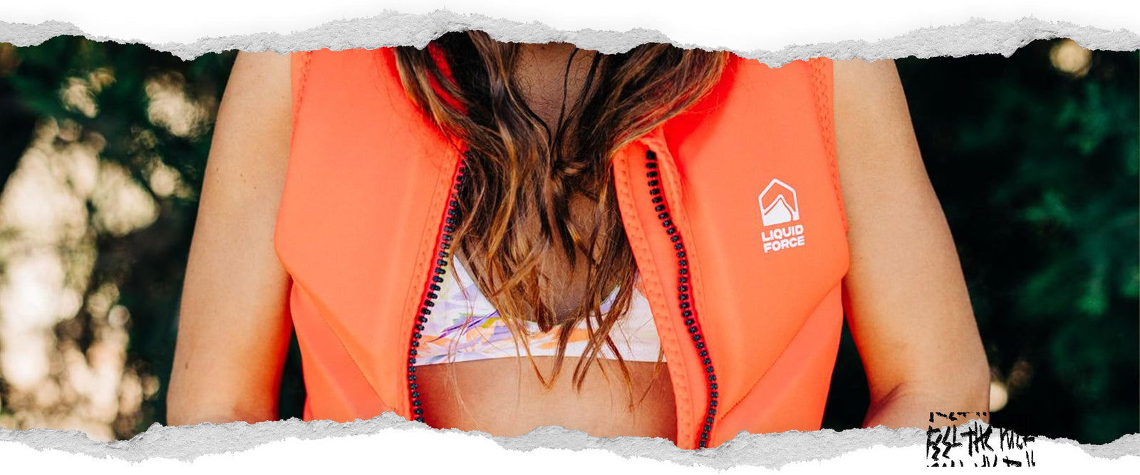 Woman in red Liquid Force vest and swimsuit zips up in front of green background.