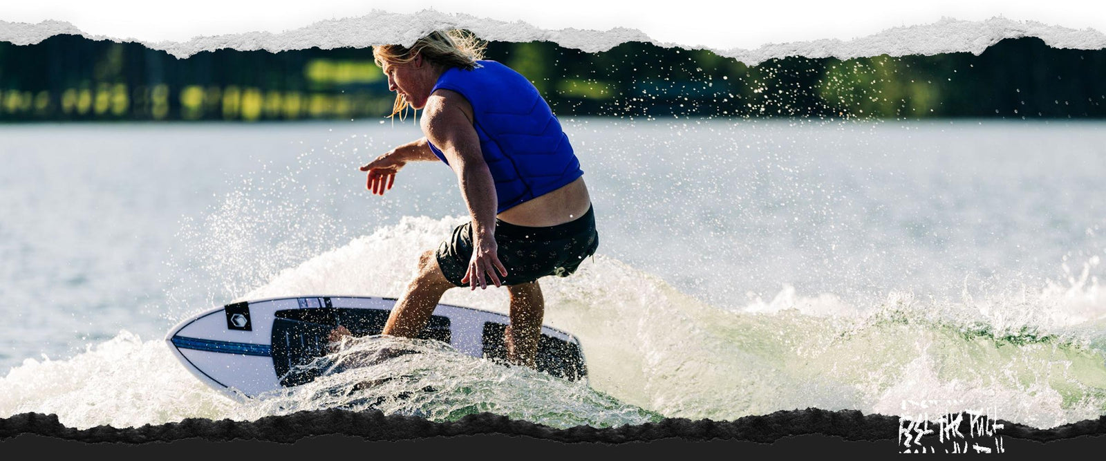 Rider in blue vest bottom turning on a white and blue wakesurf board on a lake.