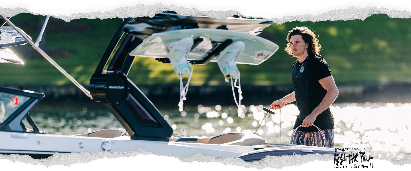 Man in black Liquid Force vest gripping a tow rope near wakeboards and a boat.