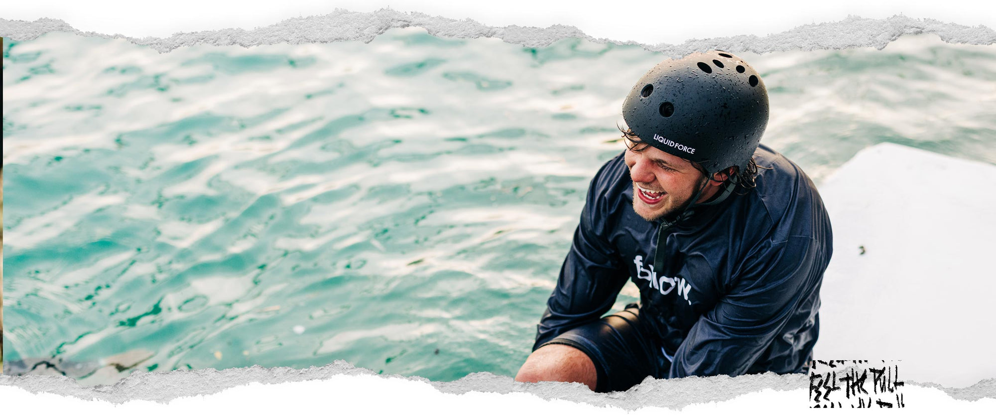 Smiling man in Liquid Force helmet sitting on a dock in wet black gear with water in background.