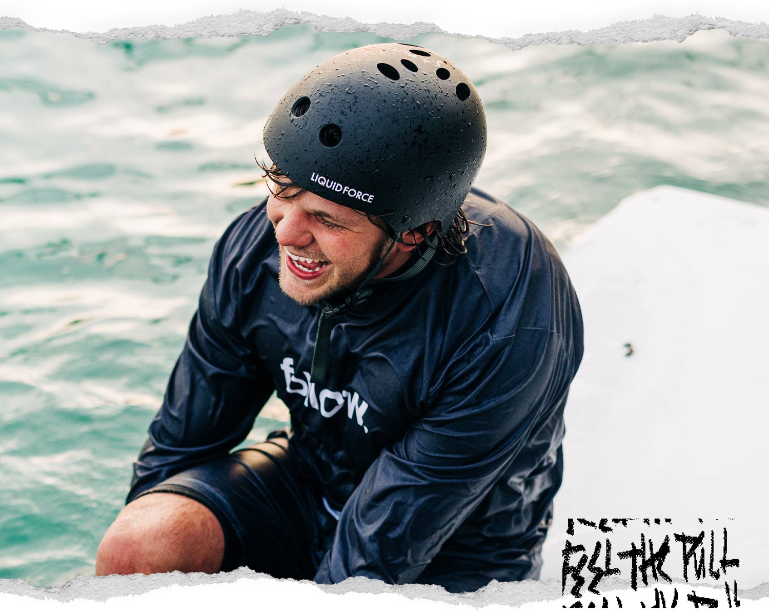 Smiling man in Liquid Force helmet sitting on a dock in wet black gear with water in background.