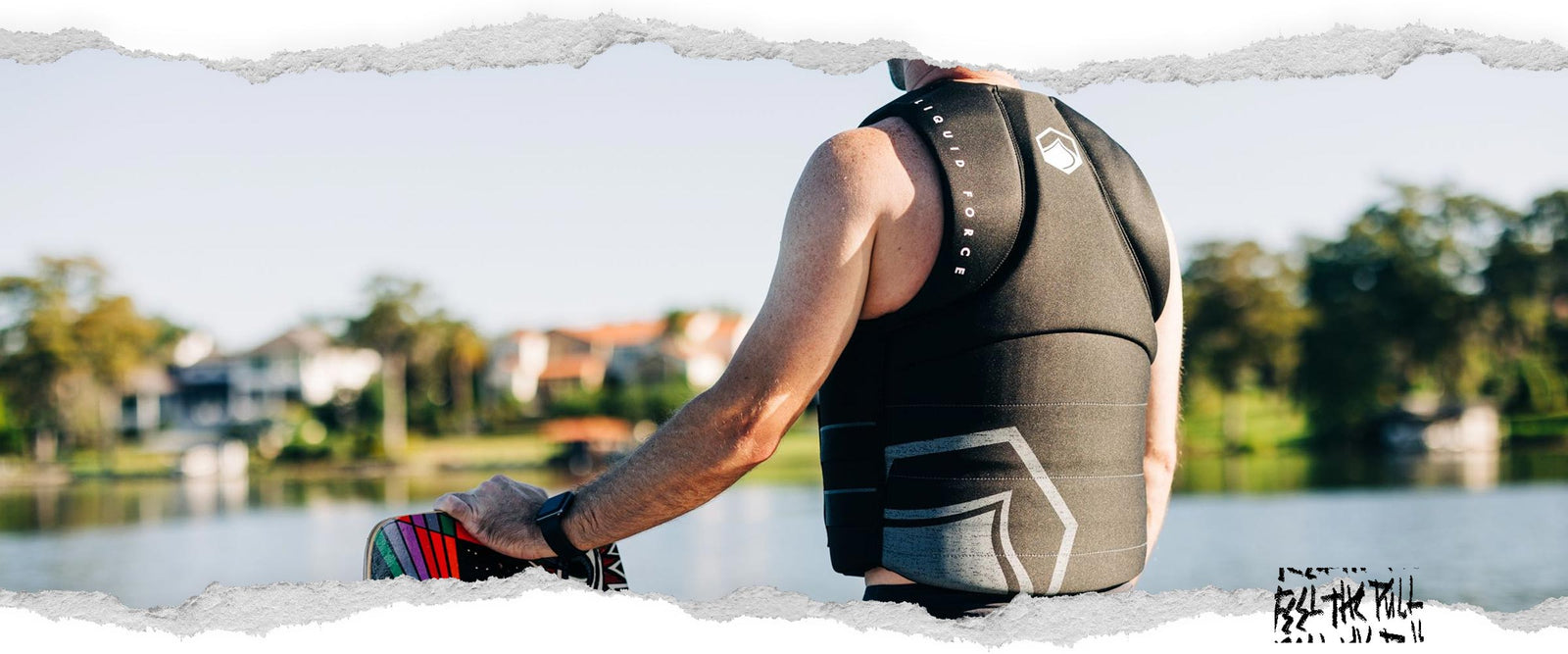 Back view of man in black Hinge CGA vest holding a wakeboard, facing lake in morning light.