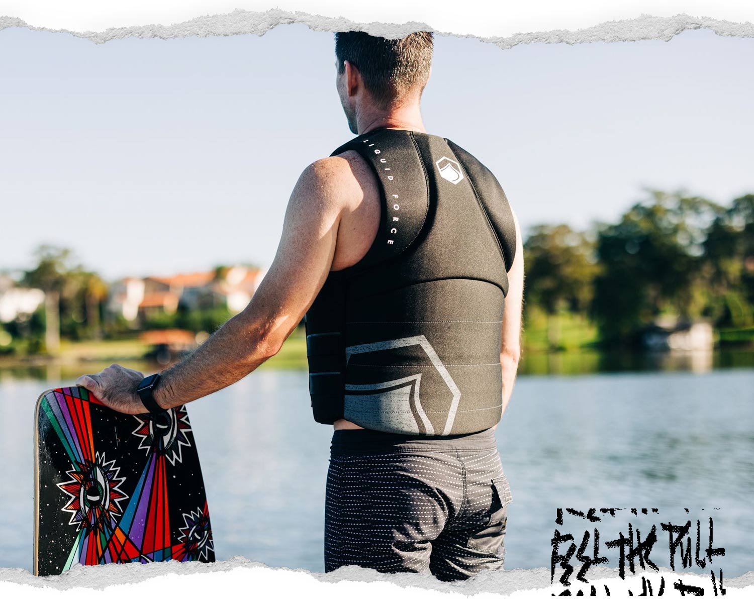 Back view of man in black Hinge CGA vest holding a wakeboard, facing lake in morning light.