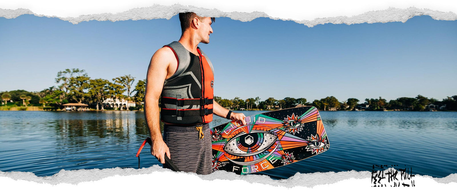 Man in red and black Hinge CGA vest stands by a lake holding a colorful wakeboard.