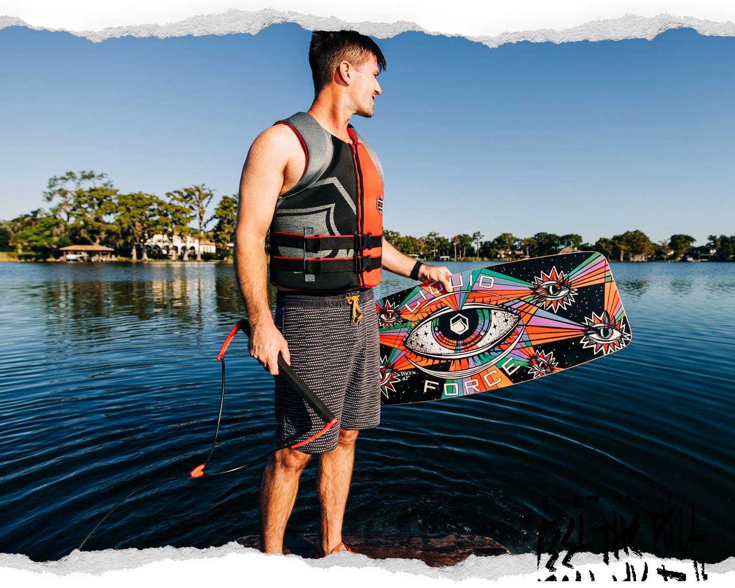 Man in red and black Hinge CGA vest stands by a lake holding a colorful wakeboard.