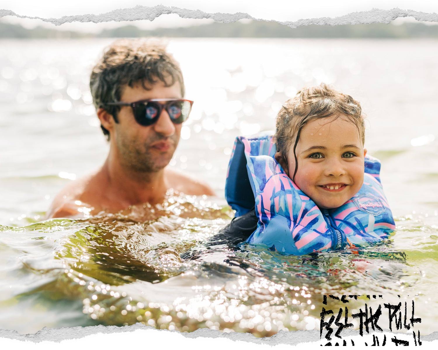 Smiling child in a tropical blue and pink CGA vest floats in water with adult behind them.