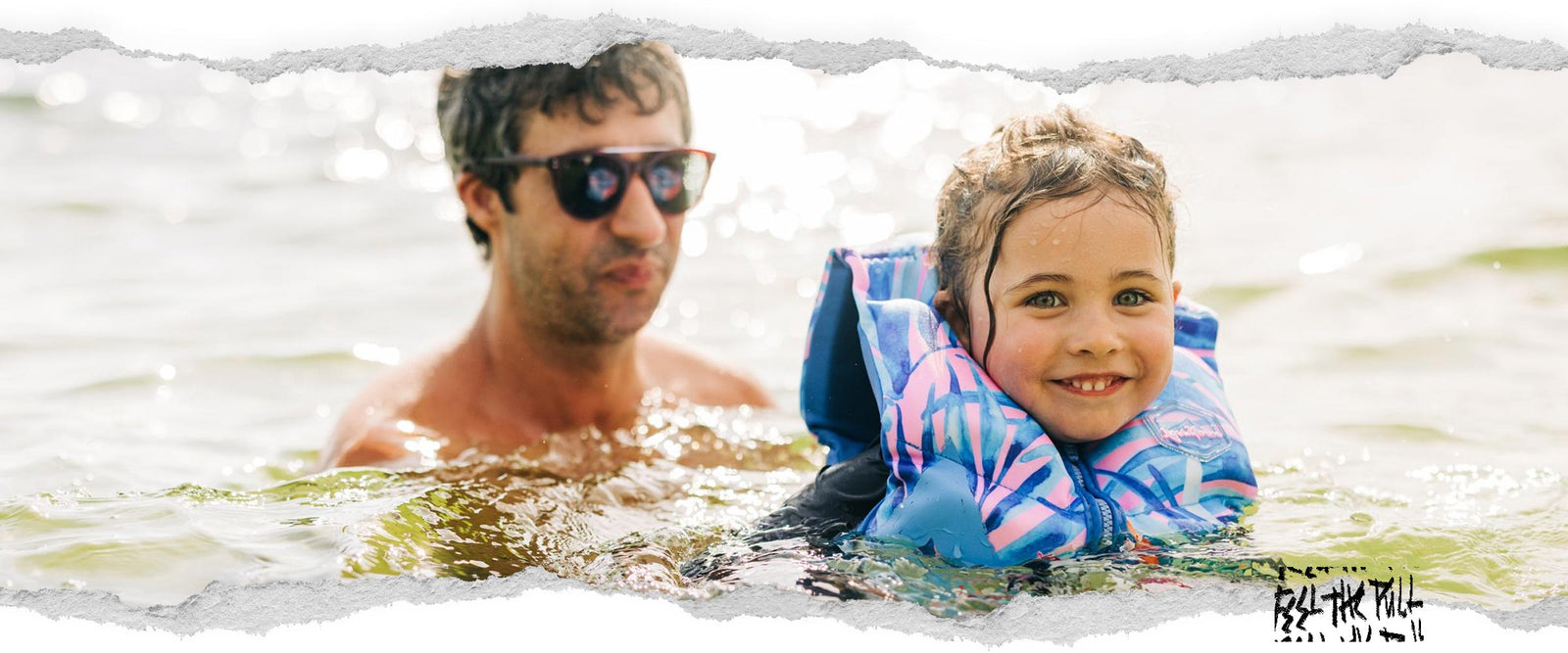 Smiling child in a tropical blue and pink CGA vest floats in water with adult behind them.