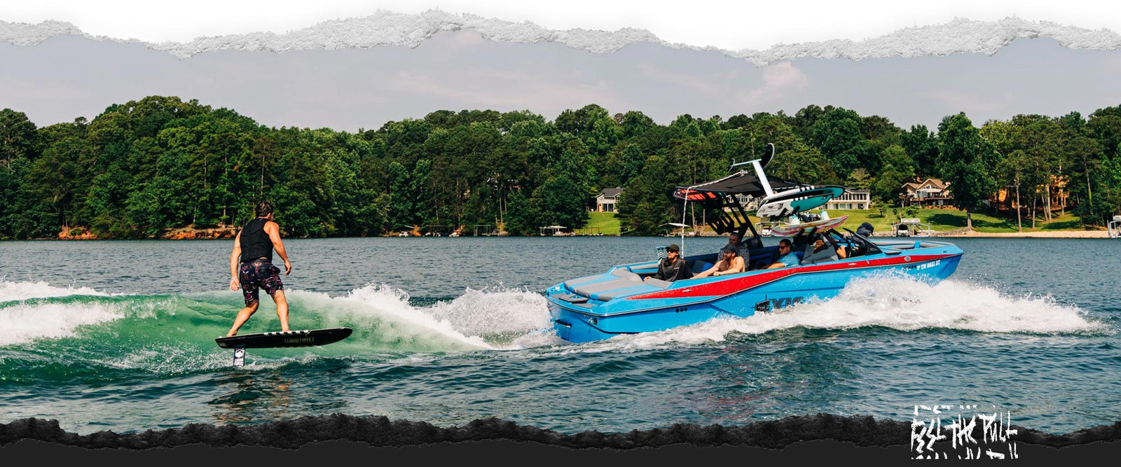 Surfer foiling behind a blue and red boat with wooded shoreline in background.