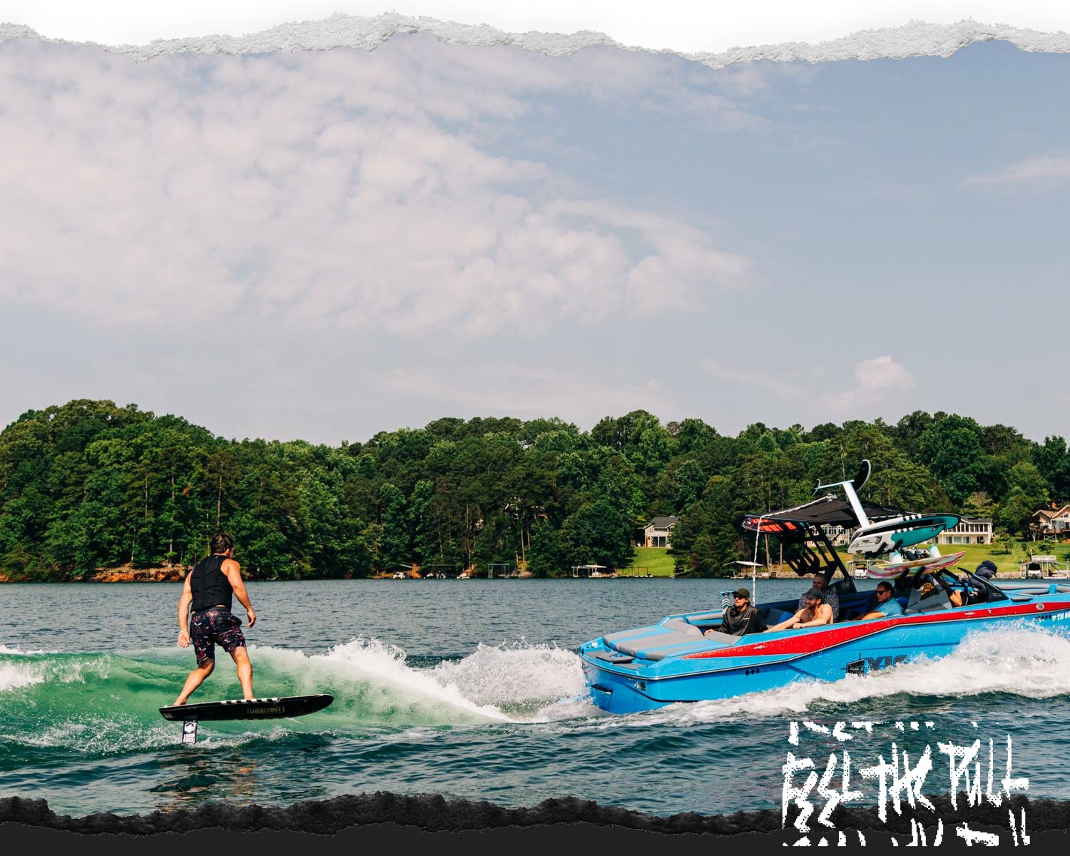 Surfer foiling behind a blue and red boat with wooded shoreline in background.