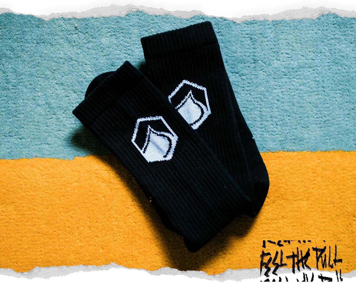 Pair of black socks with white logo on a textured teal and yellow surface.