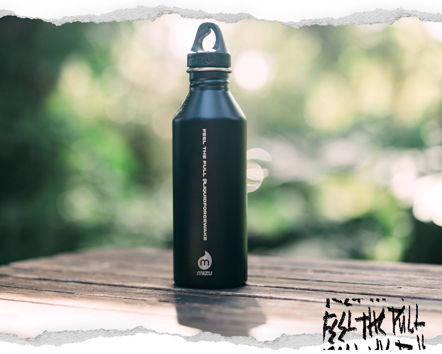 A black metal water bottle with the text "FEEL THE PULL" and blurred greenery in the background.