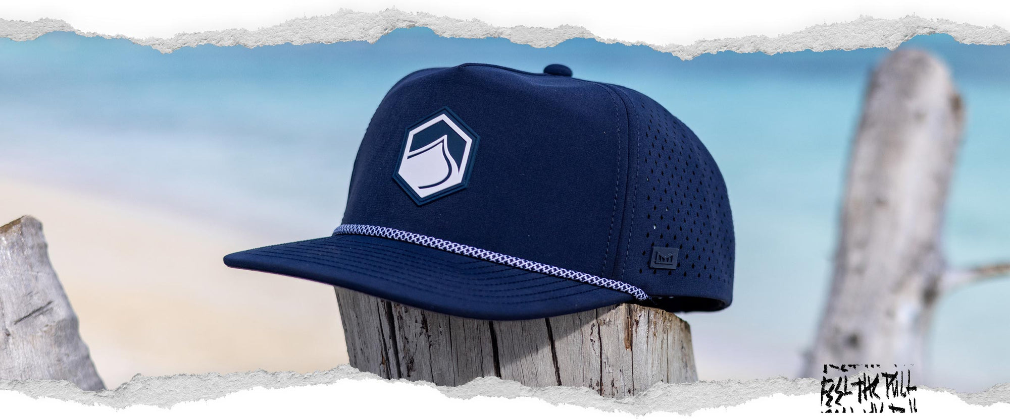 Navy flat-brim hat with hexagon wave logo and perforated sides, shown on a beachside wood post.