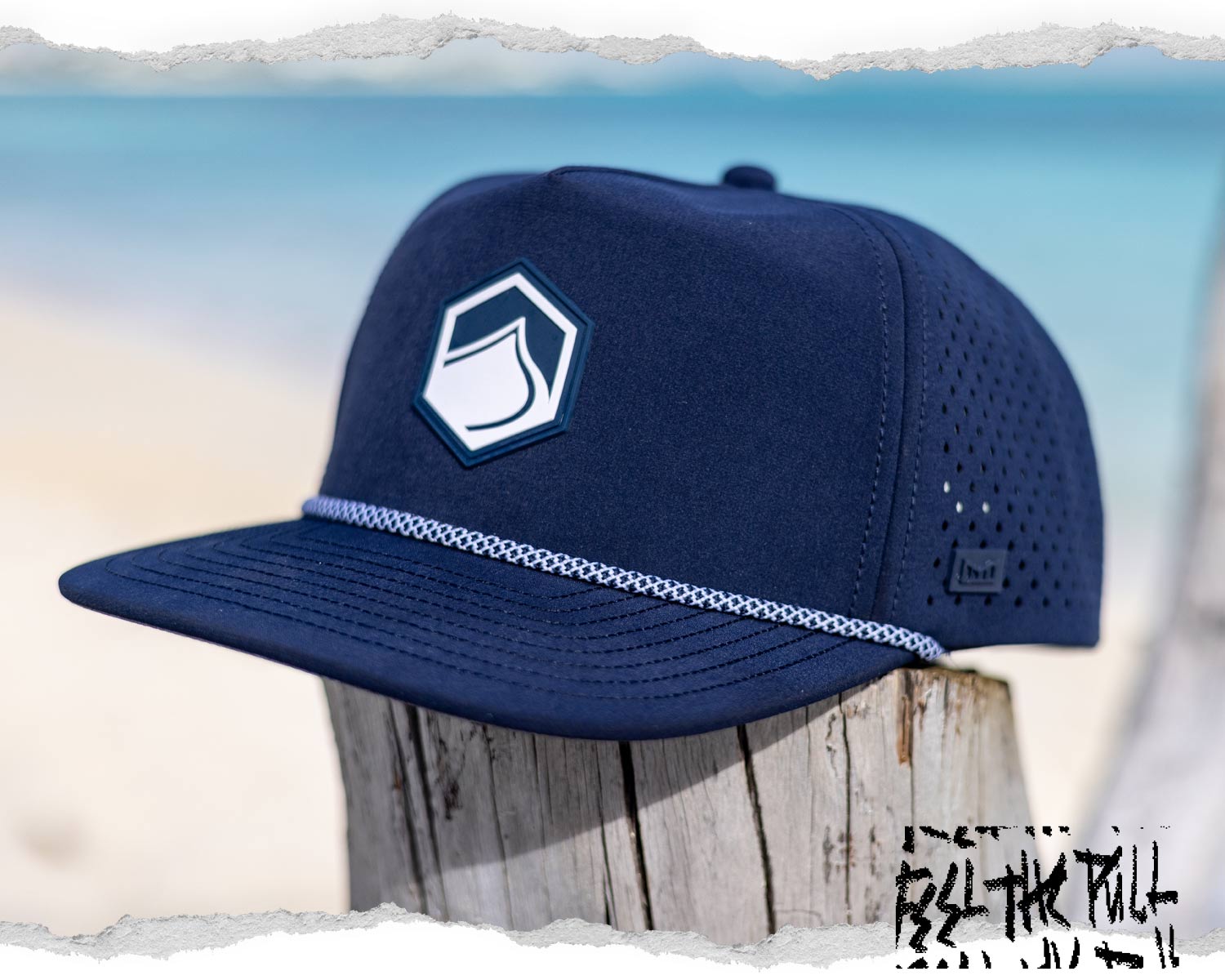 Navy flat-brim hat with hexagon wave logo and perforated sides, shown on a beachside wood post.