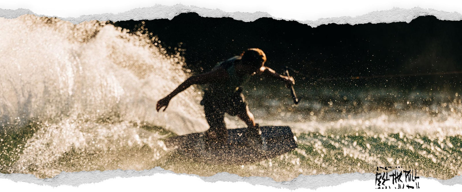 Wakeboarder carving hard with water spray lit by sunlight, silhouetted against a dark background.