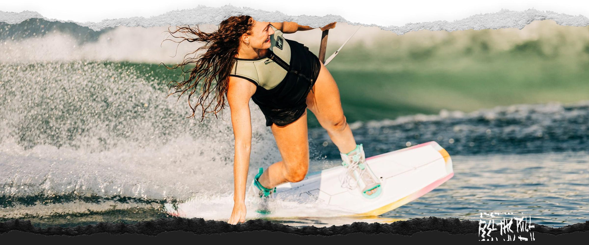 Woman wakeboarding in tan vest and teal bindings with one hand touching the water.