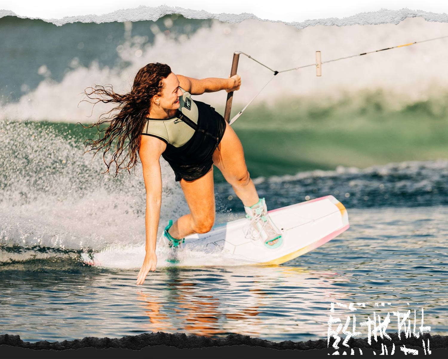 Woman wakeboarding in tan vest and teal bindings with one hand touching the water.