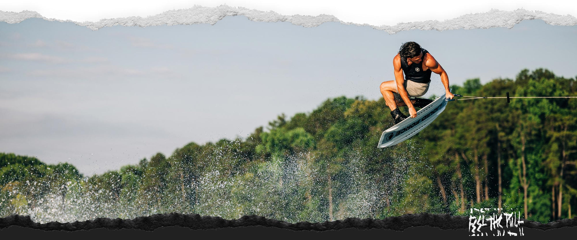 Wakeboarder grabs white Prime Aero board above water with forest in background.