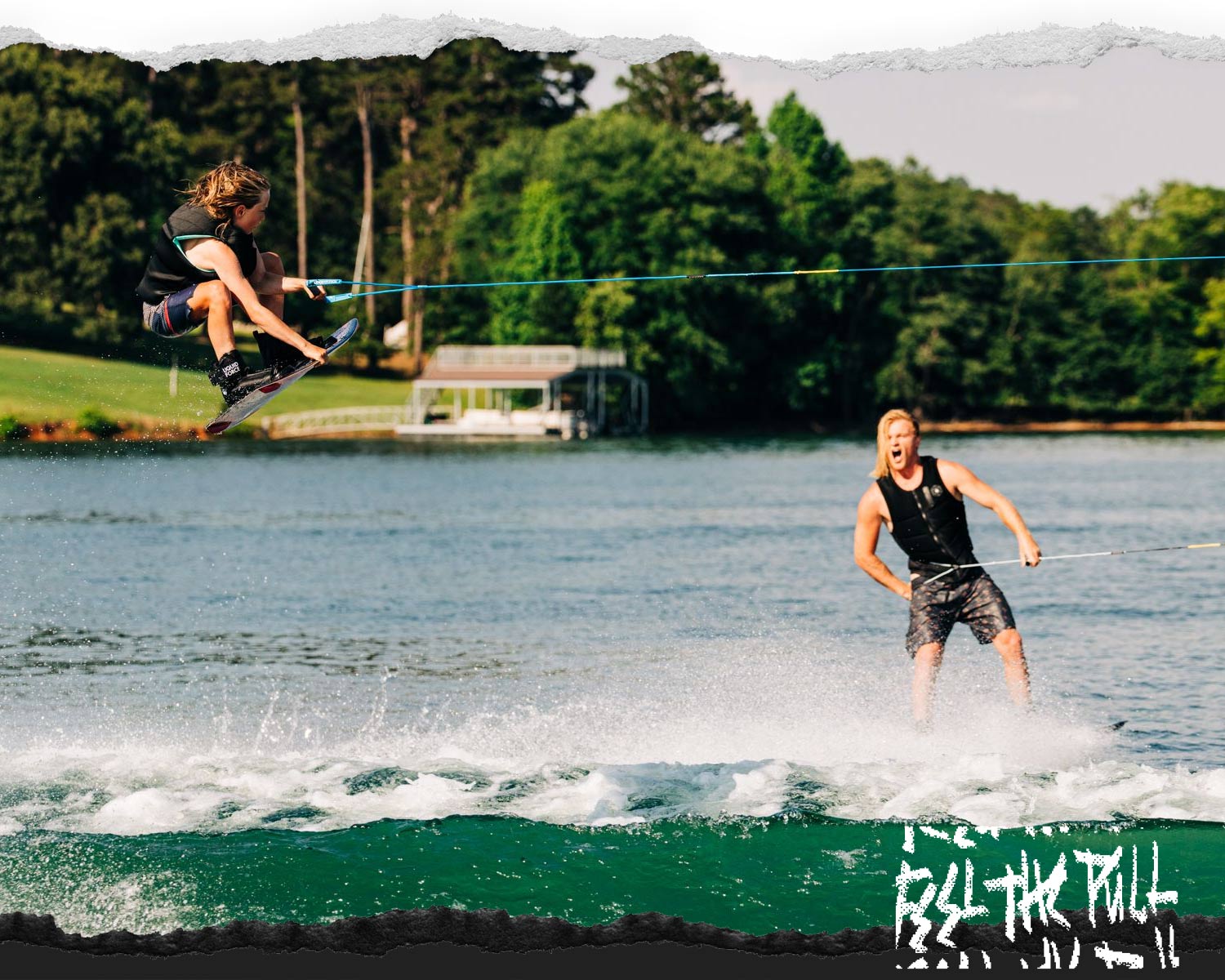 Young rider jumps while adult rides behind, both wakeboarding on a sunny lake.