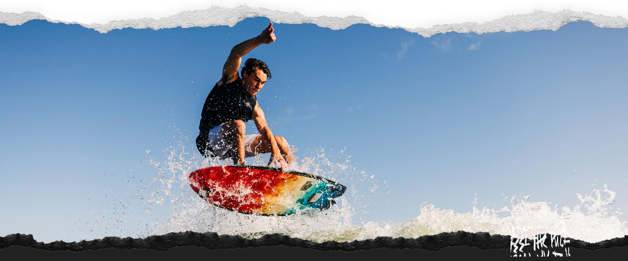 Rider launching off the wake on a multicolor wakesurf board against a clear blue sky.