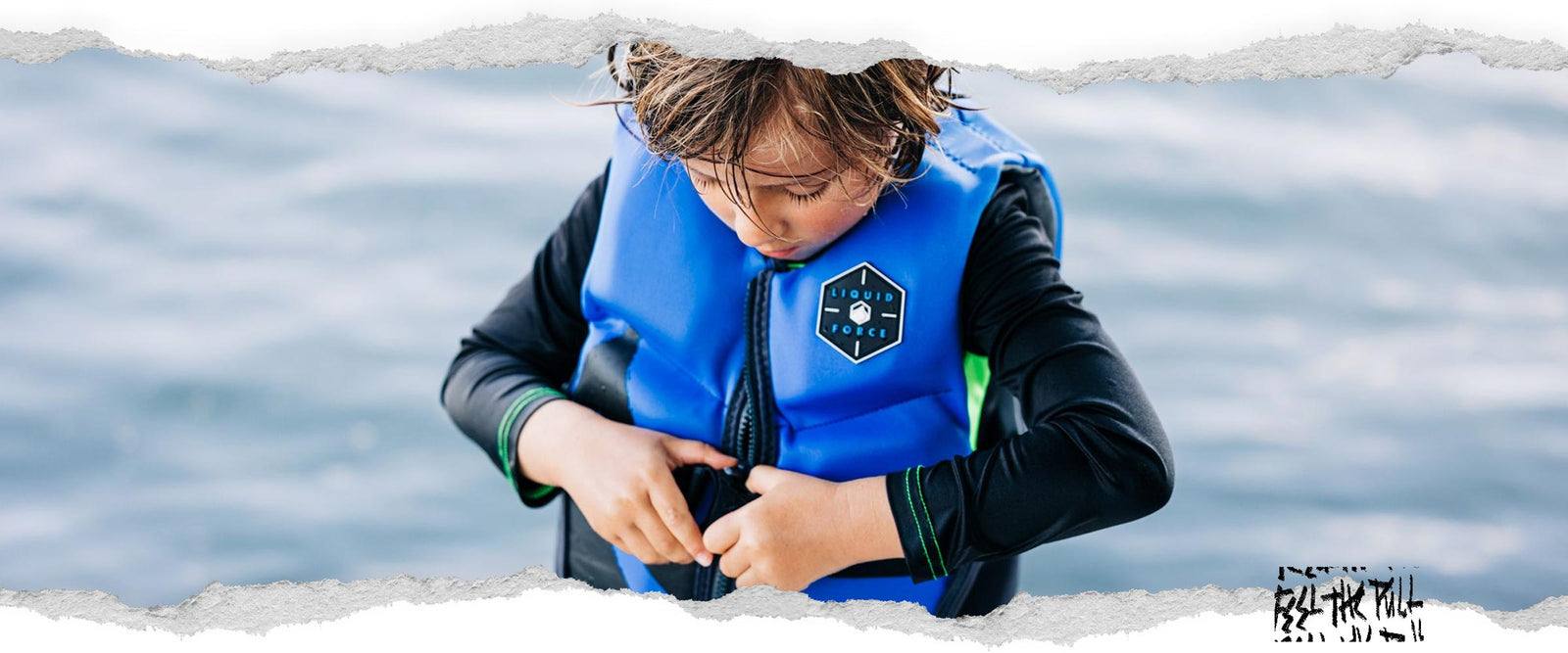 Child in blue and black Liquid Force CGA vest adjusting buckle, standing in front of water.