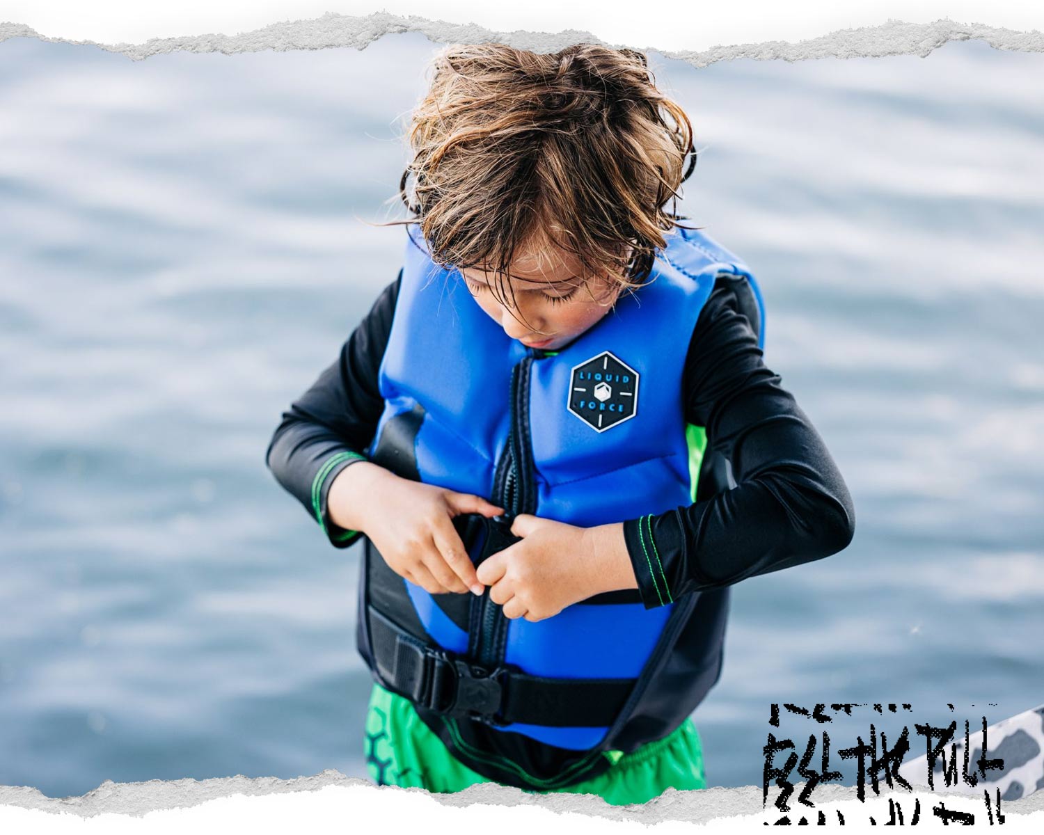 Child in blue and black Liquid Force CGA vest adjusting buckle, standing in front of water.