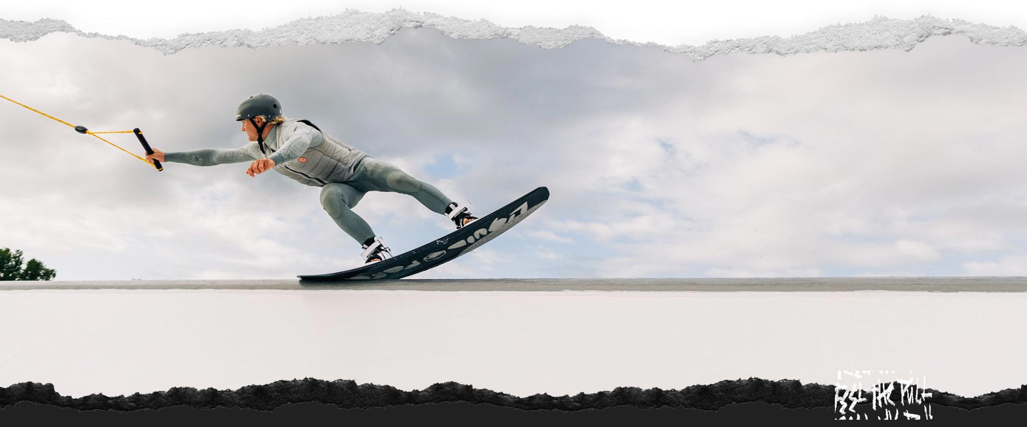 Rider in grey wetsuit and helmet grabs handle while sliding across feature.