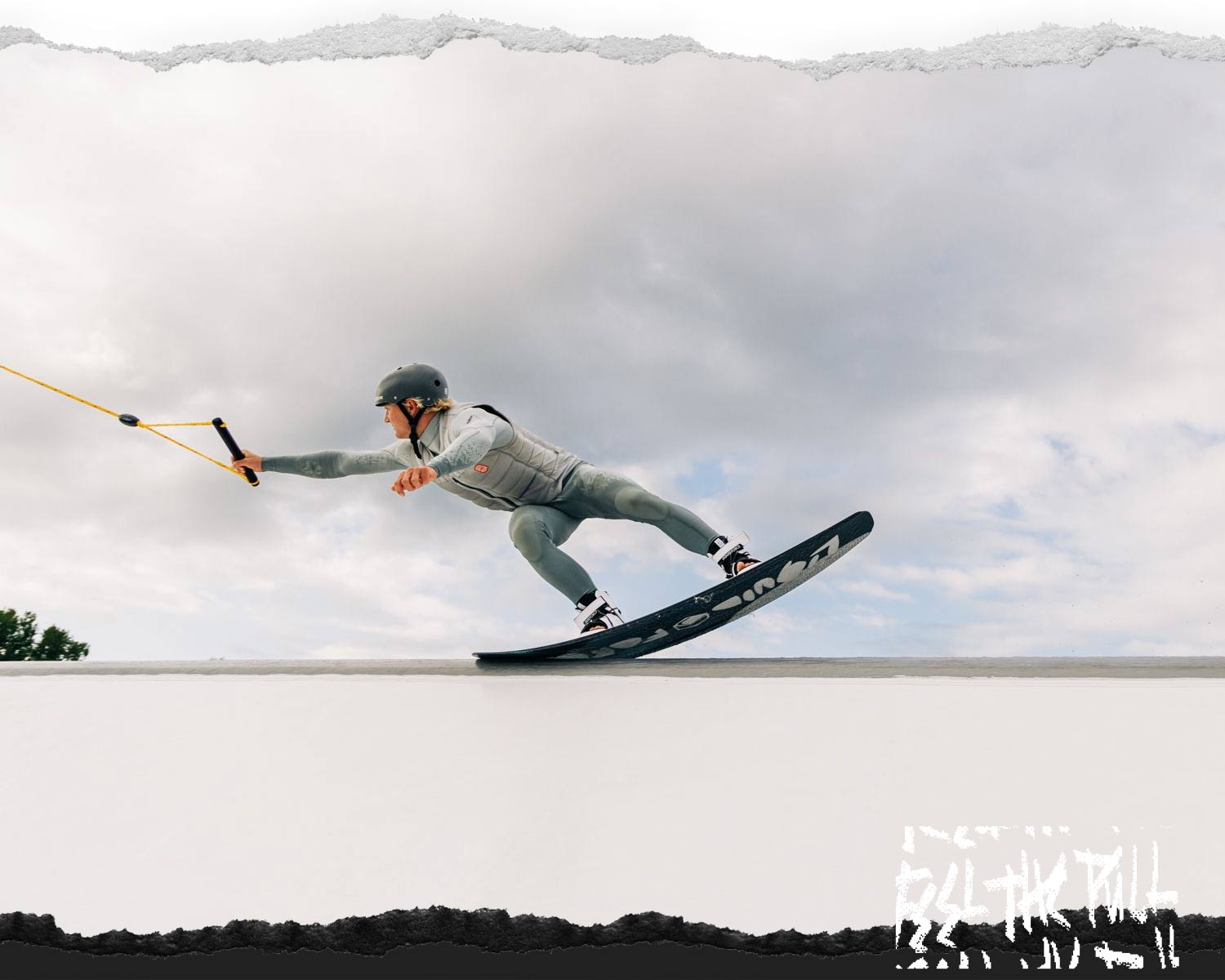 Rider in grey wetsuit and helmet grabs handle while sliding across feature.