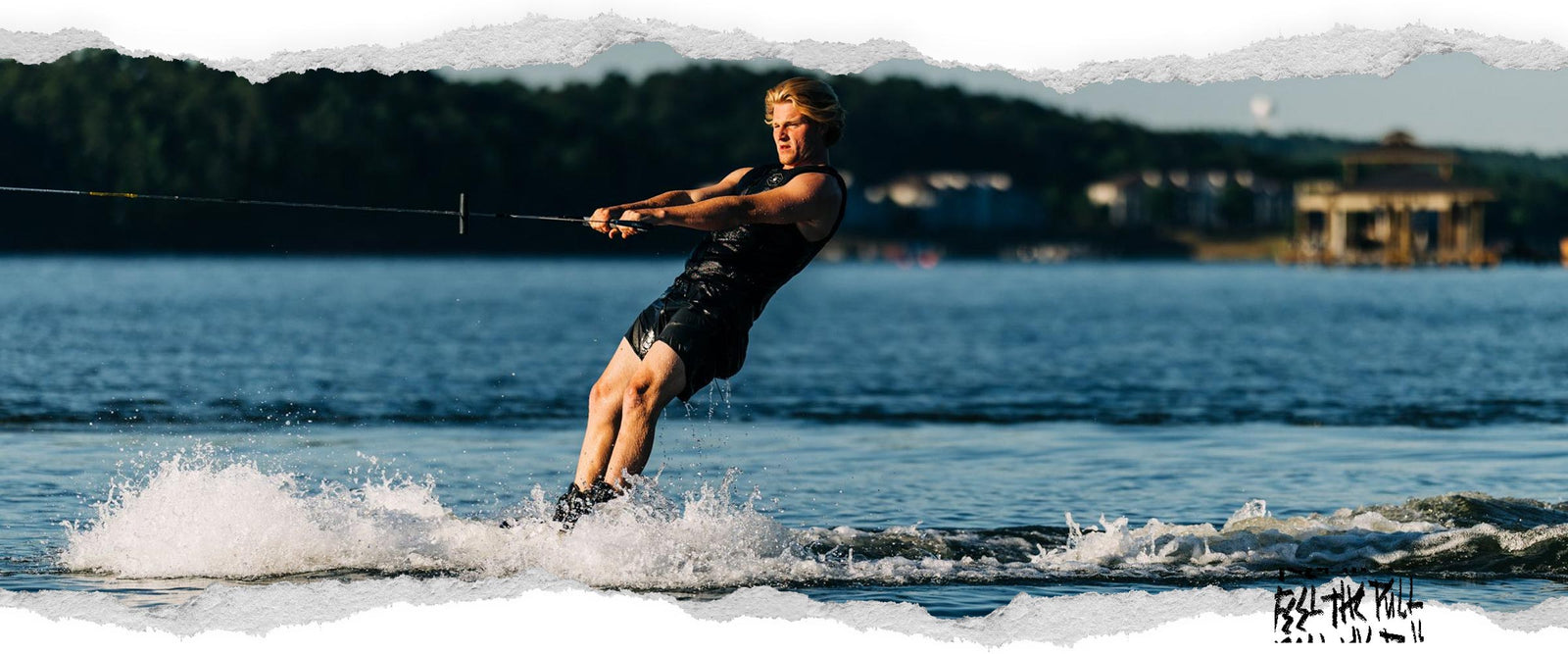 Wakeboarder skimming across water in black vest, pulling on a tow handle.