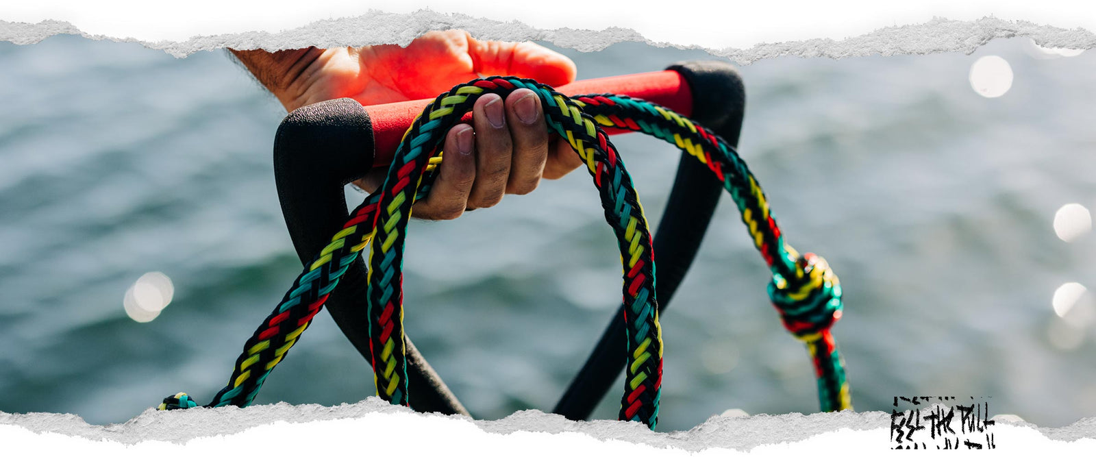 Hand holding red Liquid Force handle with rasta-colored braided rope.