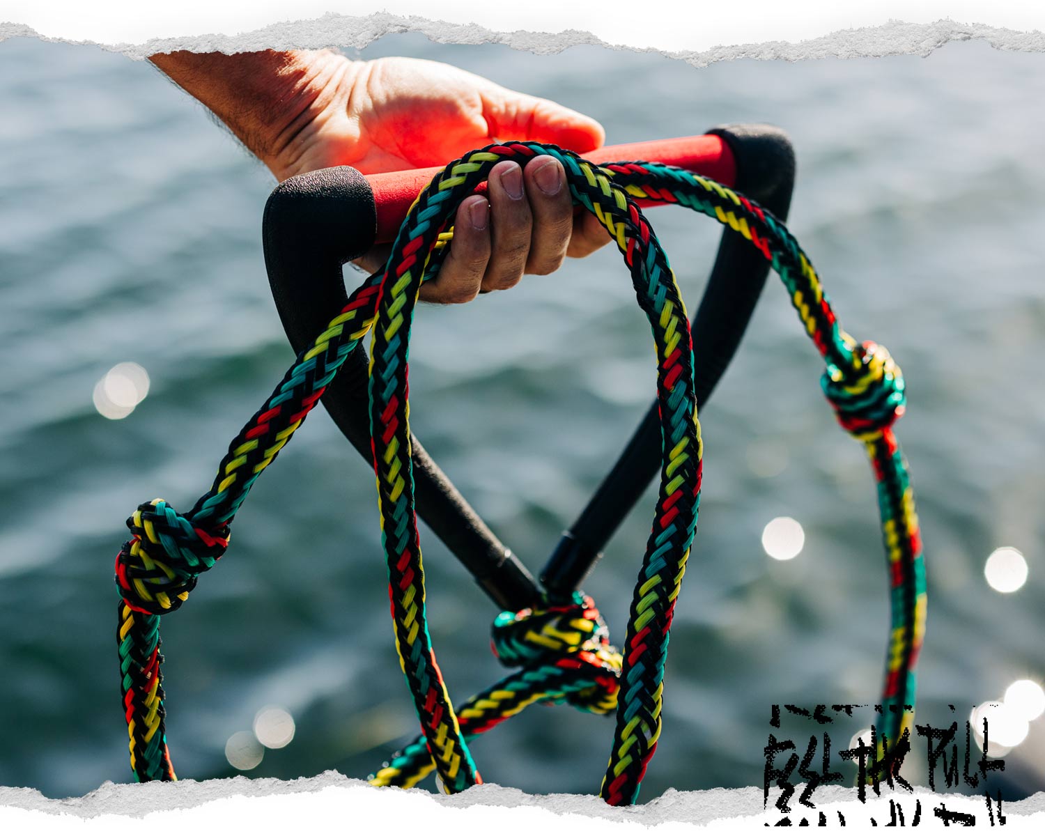 Hand holding red Liquid Force handle with rasta-colored braided rope.