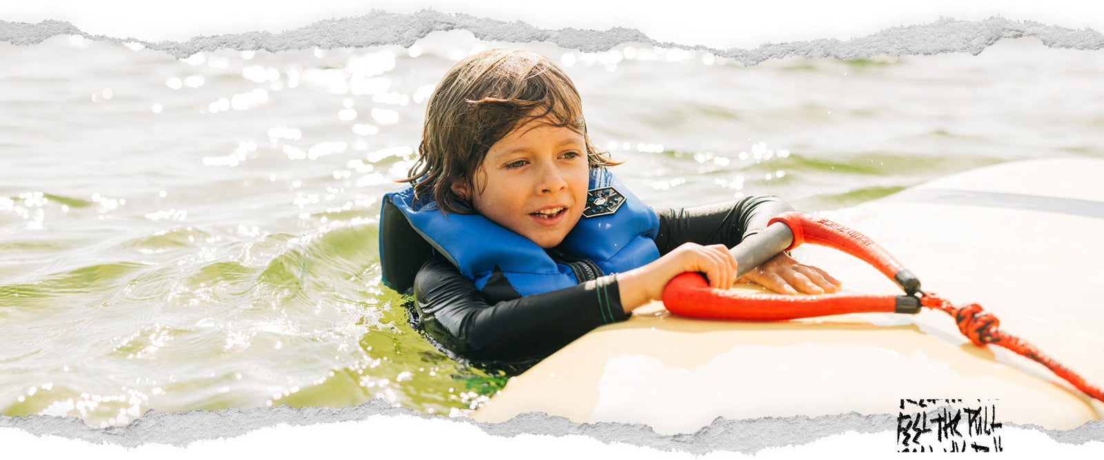 Child in blue vest holds red Liquid Force handle while floating on board.