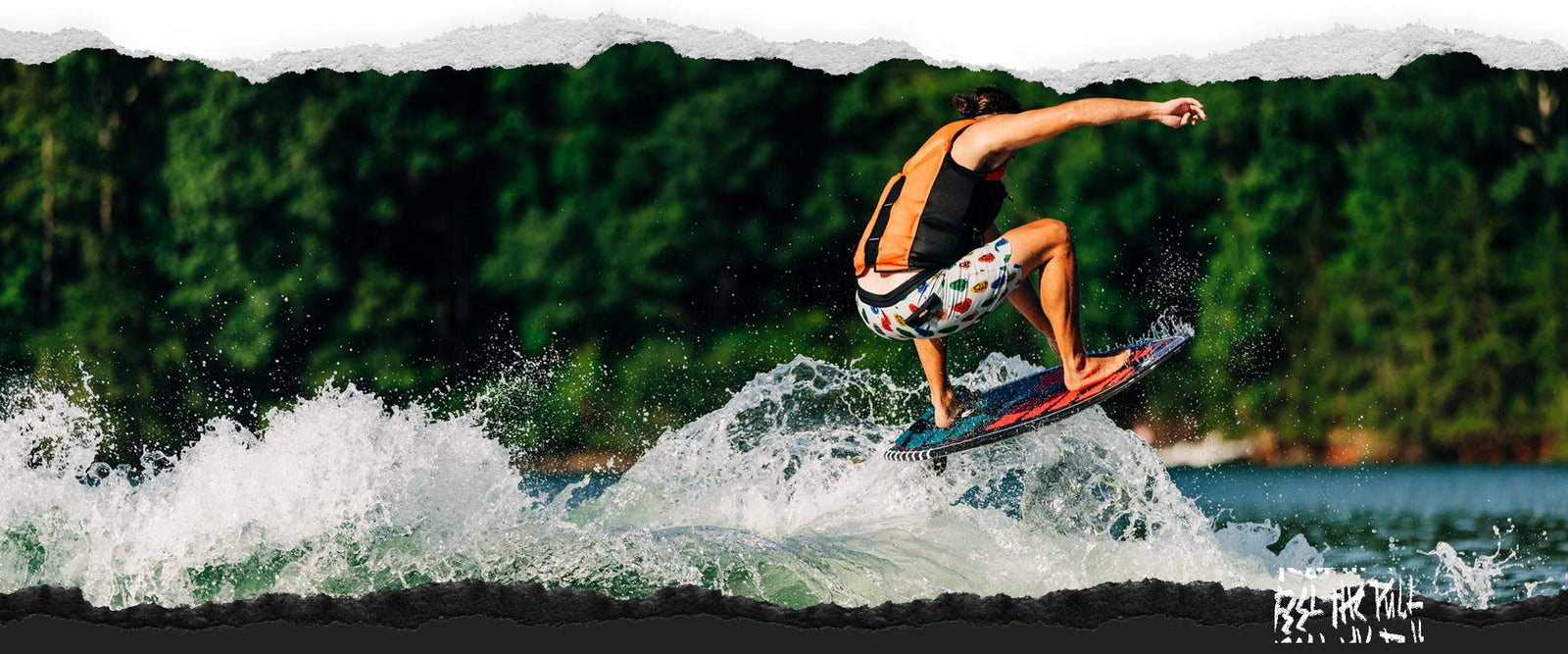 Rider in orange and black vest catching air on a red wakesurf board in front of trees.