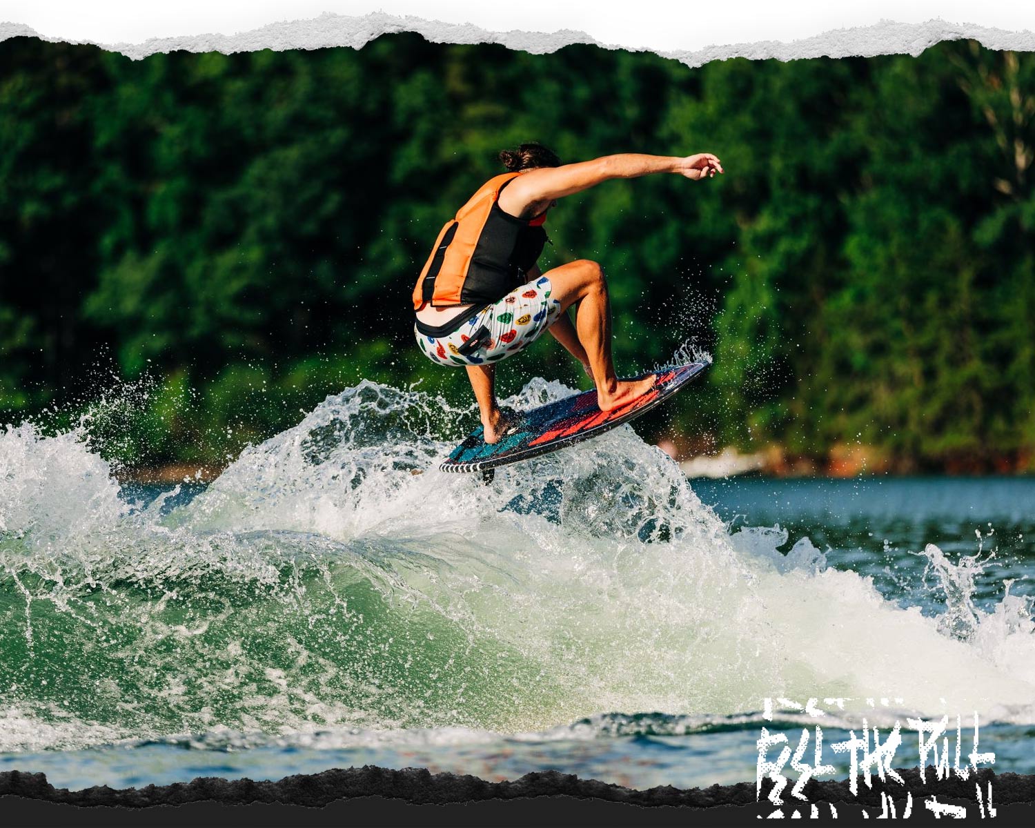 Rider in orange and black vest catching air on a red wakesurf board in front of trees.
