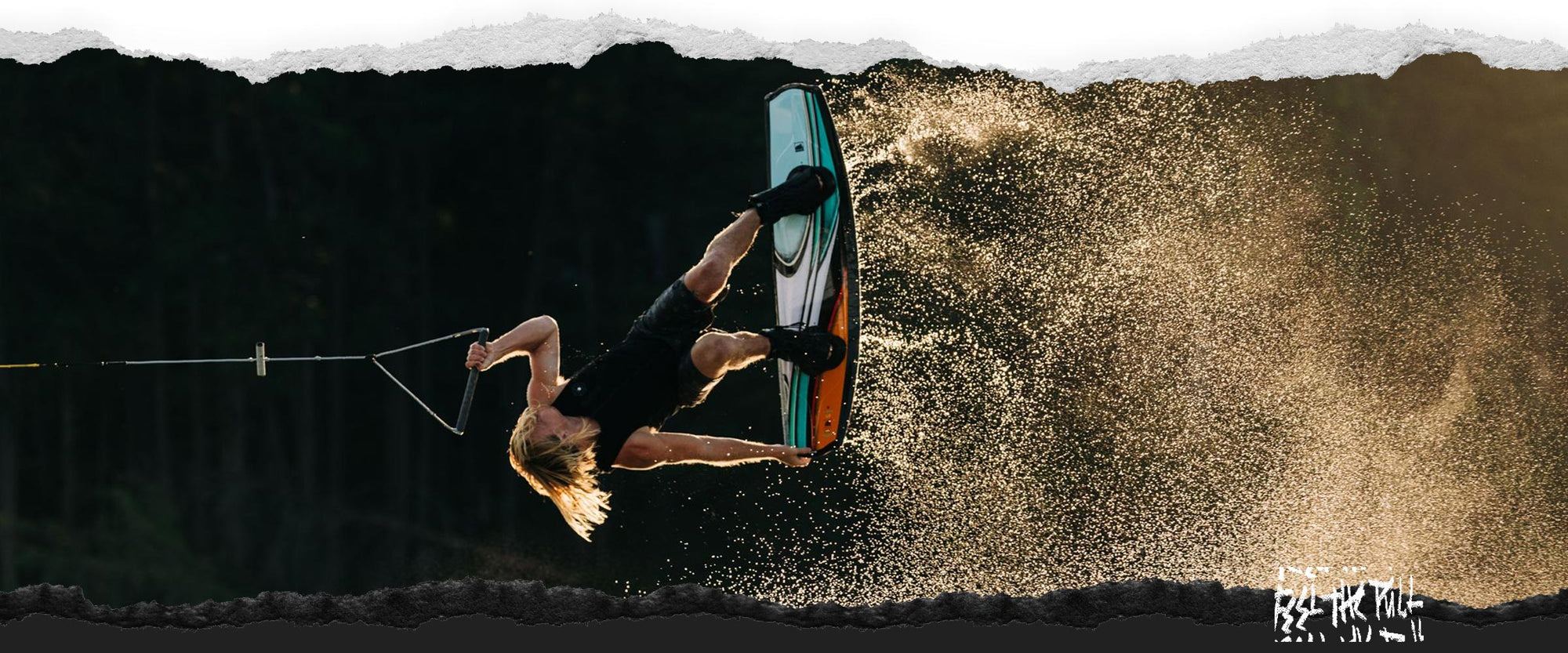 Rider doing a wakeboard flip over the water with spray lit by sunset light.