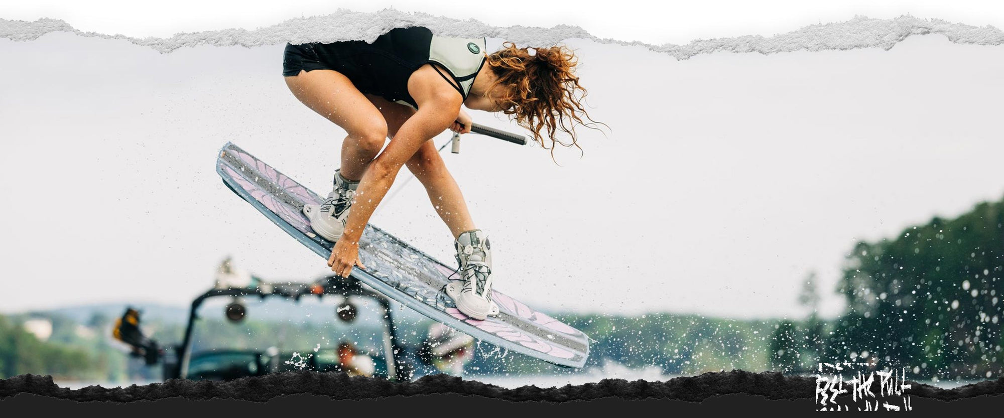 Woman in tan vest grabs wakeboard midair while being towed behind a boat.